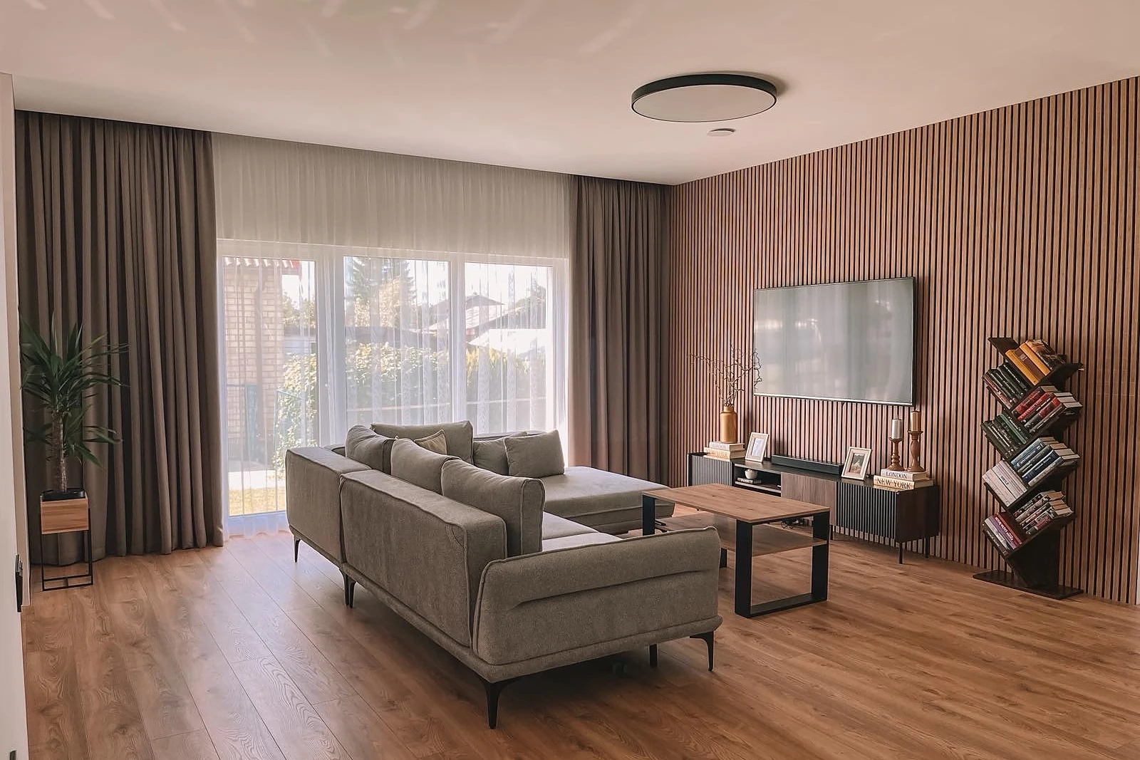 Svetainė su medinių lentelių siena, sofa ir dideliu langu po apdailos darbų